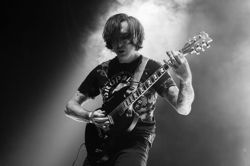 Black and white photo of a guitarist with shaggy hair playing an electric guitar on stage, surrounded by hazy smoke and dramatic lighting.