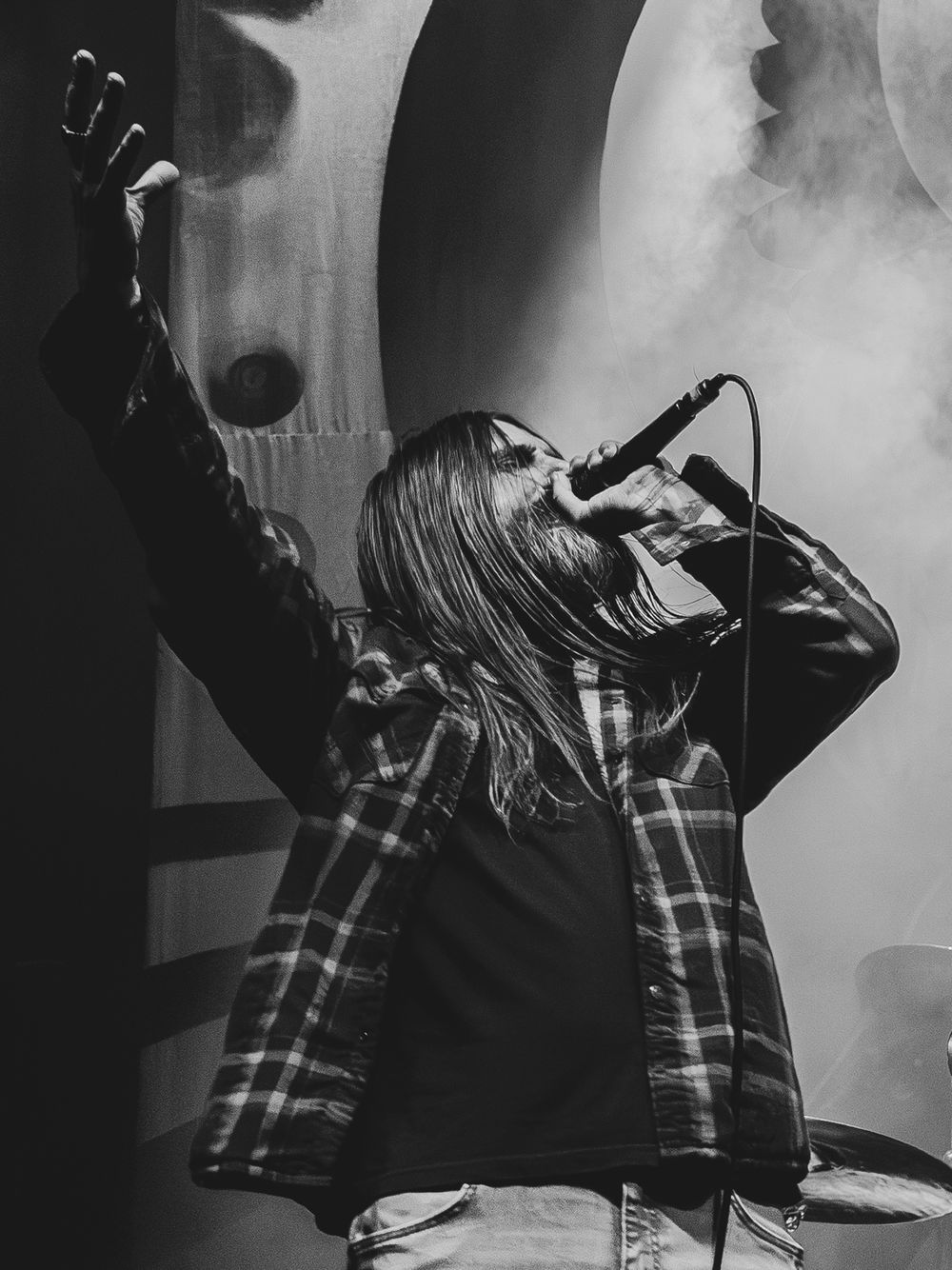 Black and white photo of a long-haired singer in a plaid flannel shirt performing live on stage with a raised arm and microphone.
