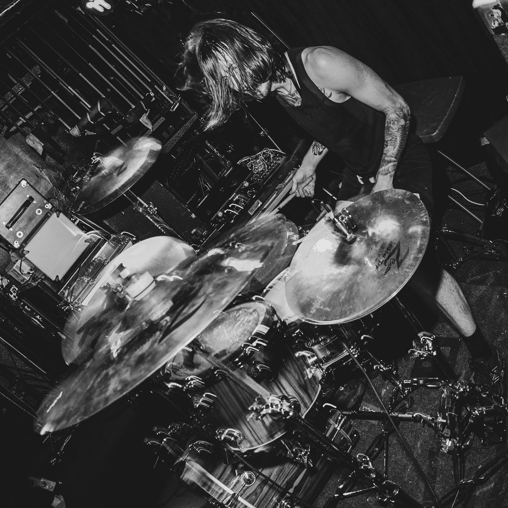 Black and white photo of a drummer performing intensely on stage with cymbals mid-motion at a live concert.