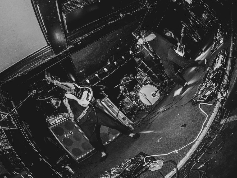Black and white aerial shot of a rock band performing on stage with guitarist, bassist, and drummer under dramatic lighting.