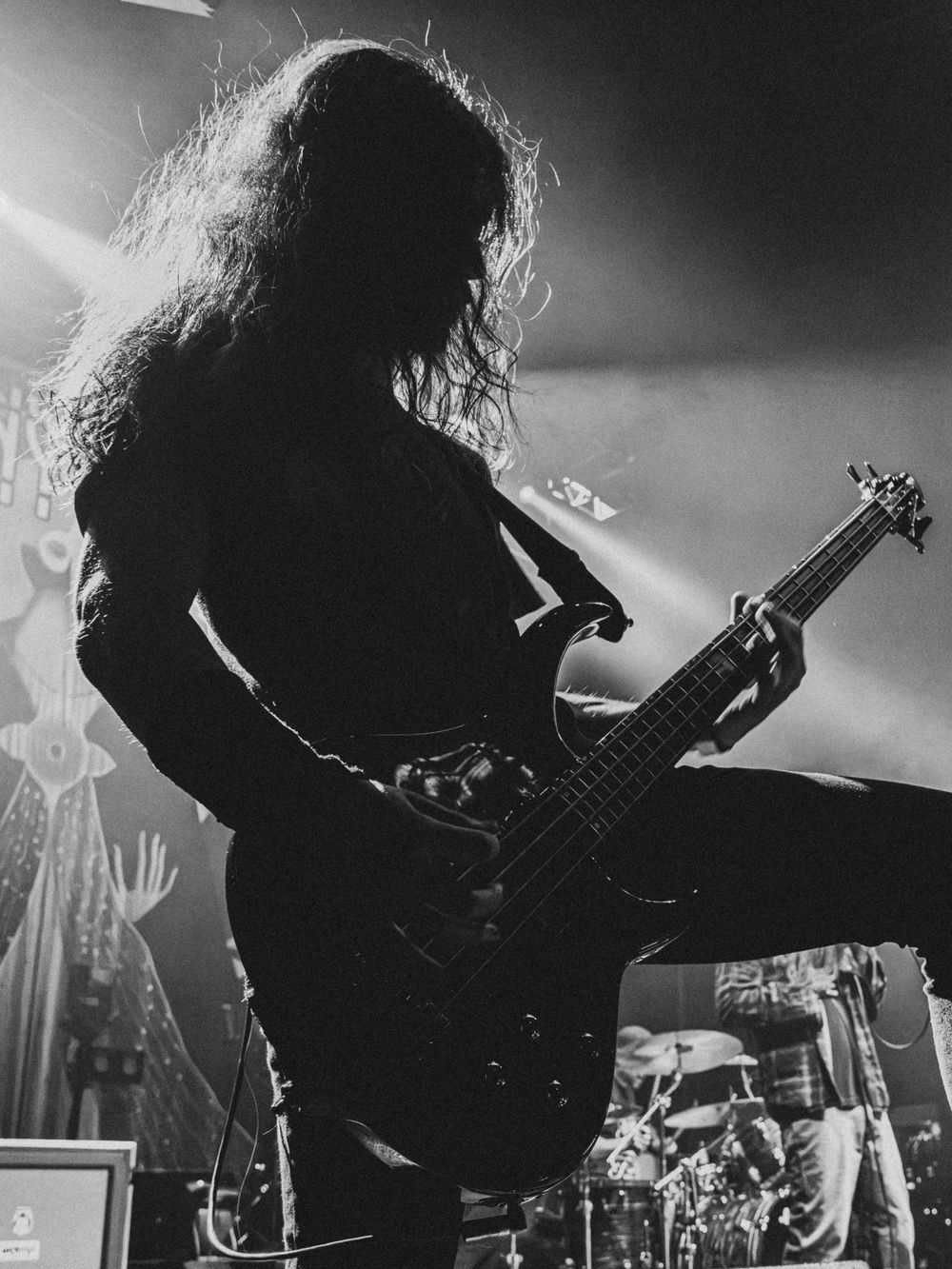 Silhouetted long-haired bassist performs on stage in black and white, with a drum kit and stage lights glowing behind.
