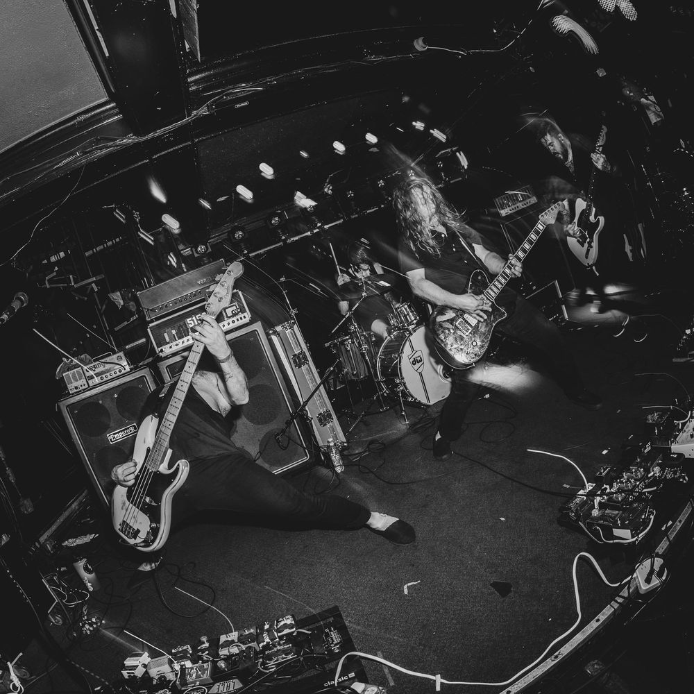 Black and white photo of a rock band performing on stage with electric guitars and amplifiers under dramatic lighting.