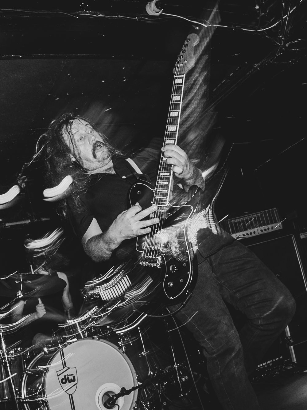 Black and white photo of a metal guitarist headbanging intensely on stage with a dark electric guitar during a live concert.