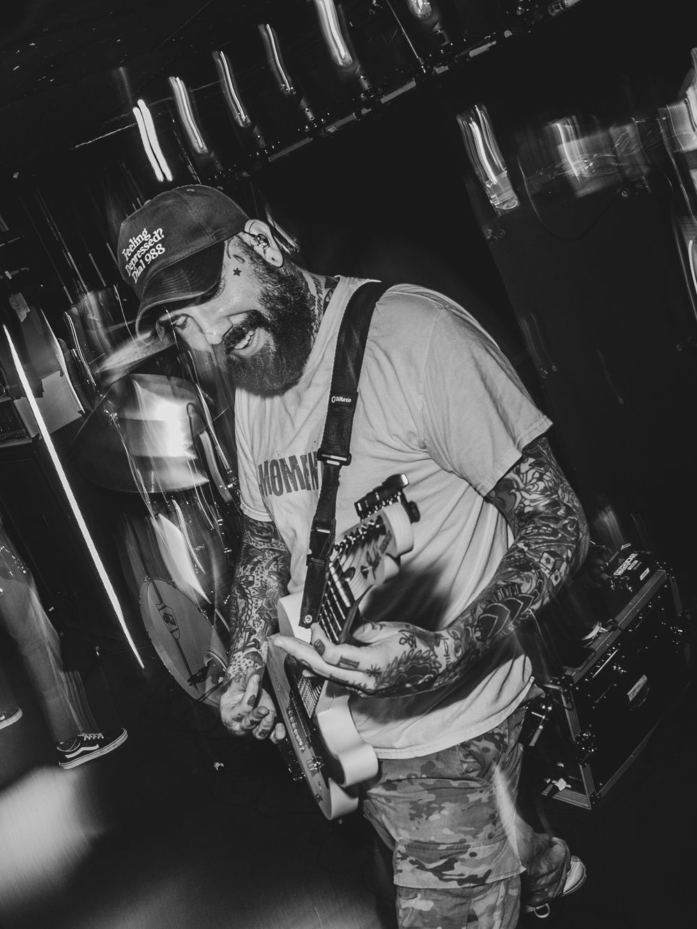 Black and white photo of a tattooed bearded man playing electric guitar on stage wearing a cap.