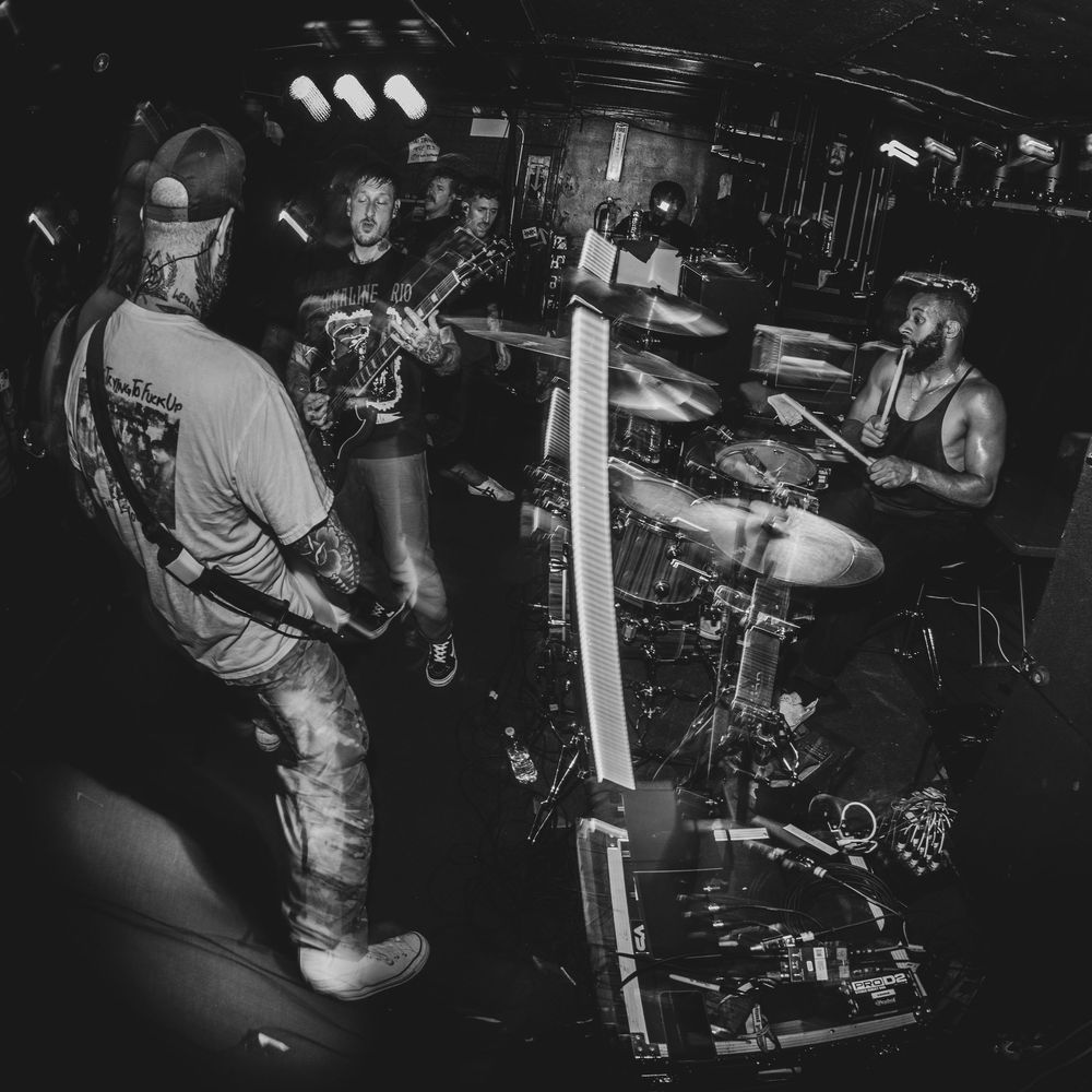 Black and white photo of a rock band performing live, with guitarist, drummer, and vocalist on a small club stage.