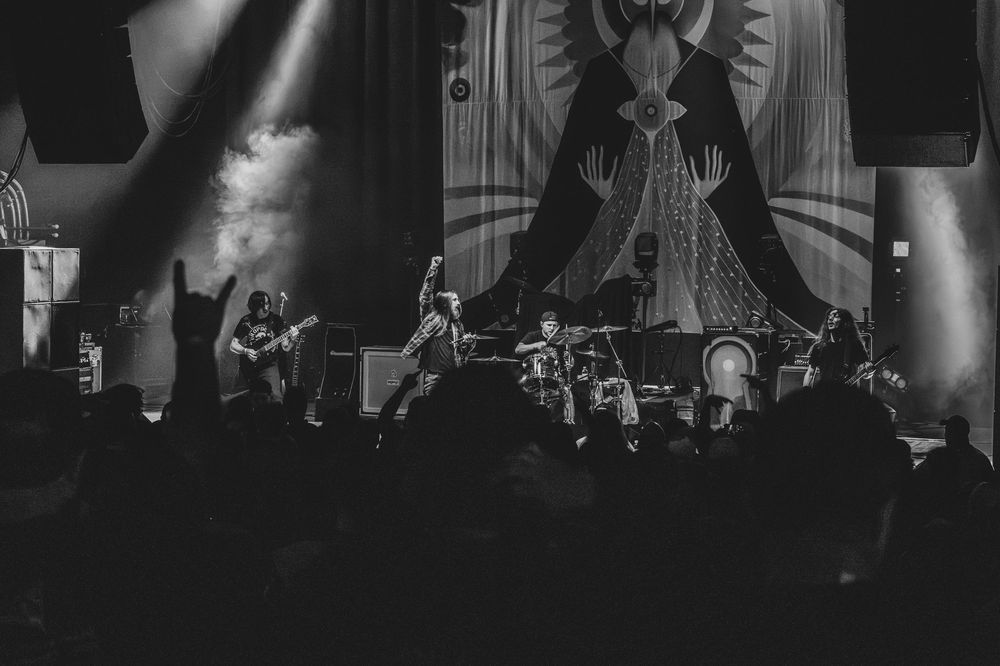 Black and white photo of a band performing live on a smoky stage before a large ornate backdrop, with a silhouetted crowd in the foreground.