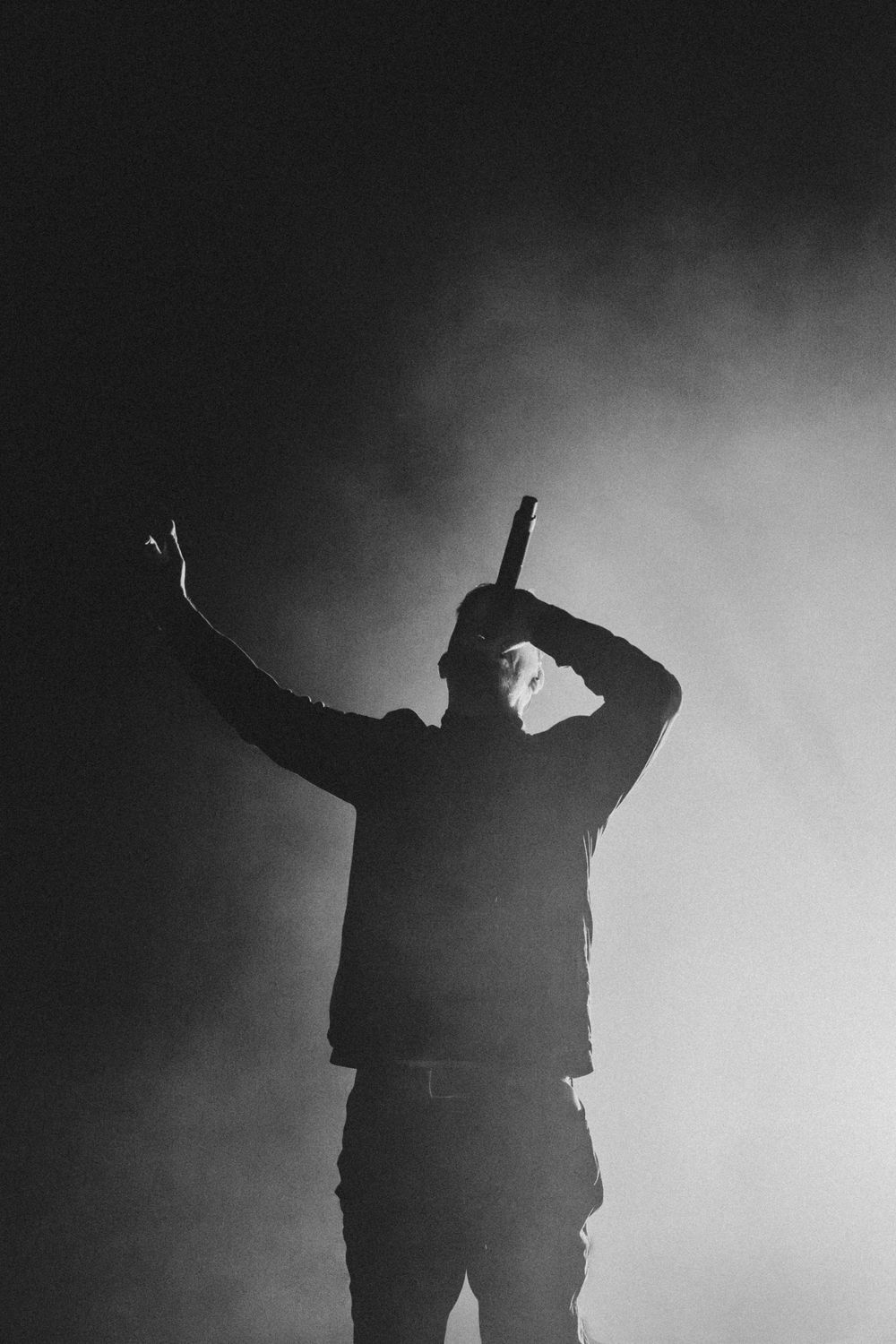 Black and white silhouette of a performer on stage holding a microphone with one arm raised, surrounded by dramatic smoke and backlighting.