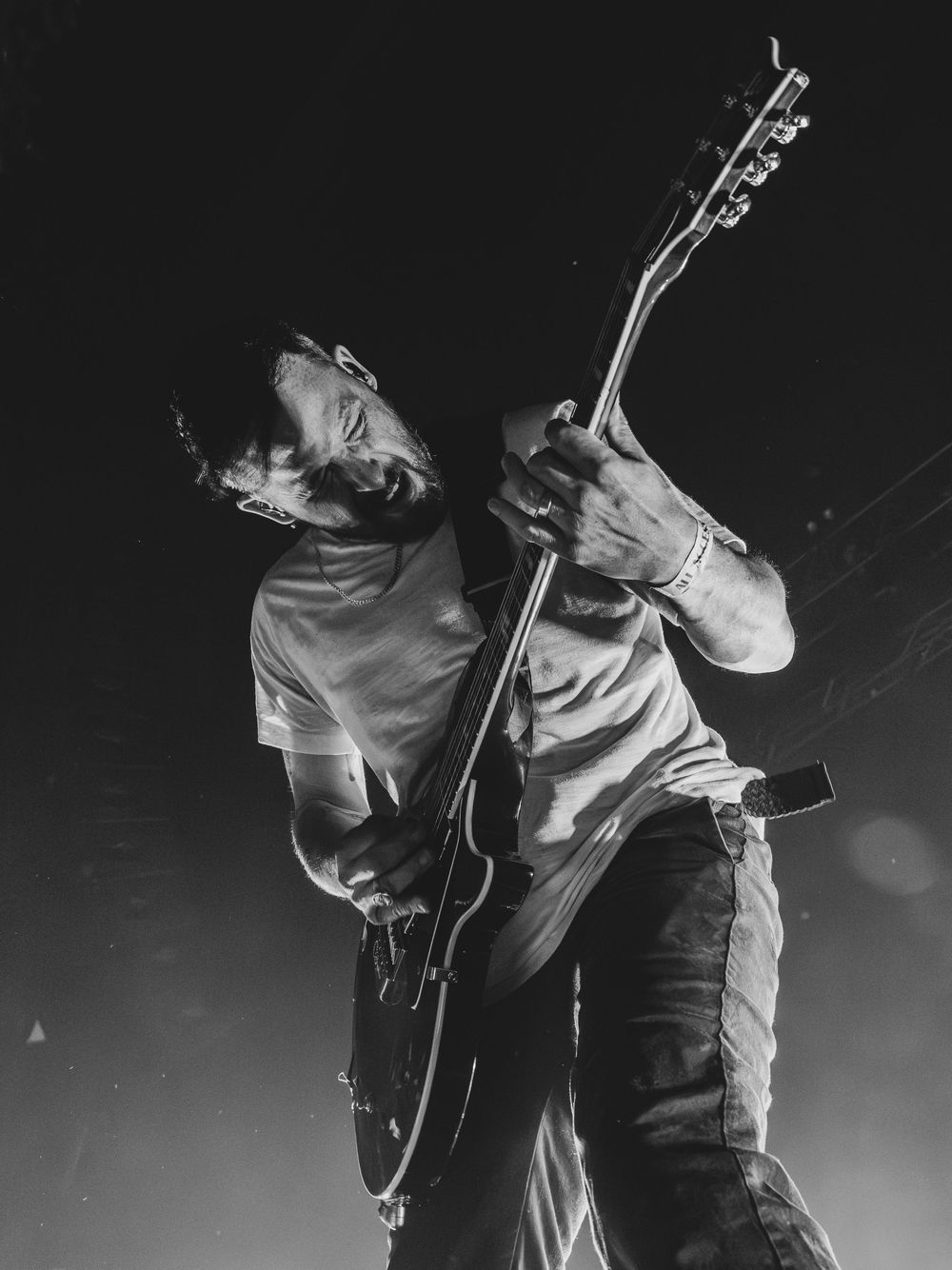 Black and white photo of a bearded guitarist leaning back while playing an electric guitar on a dark stage during a live concert.