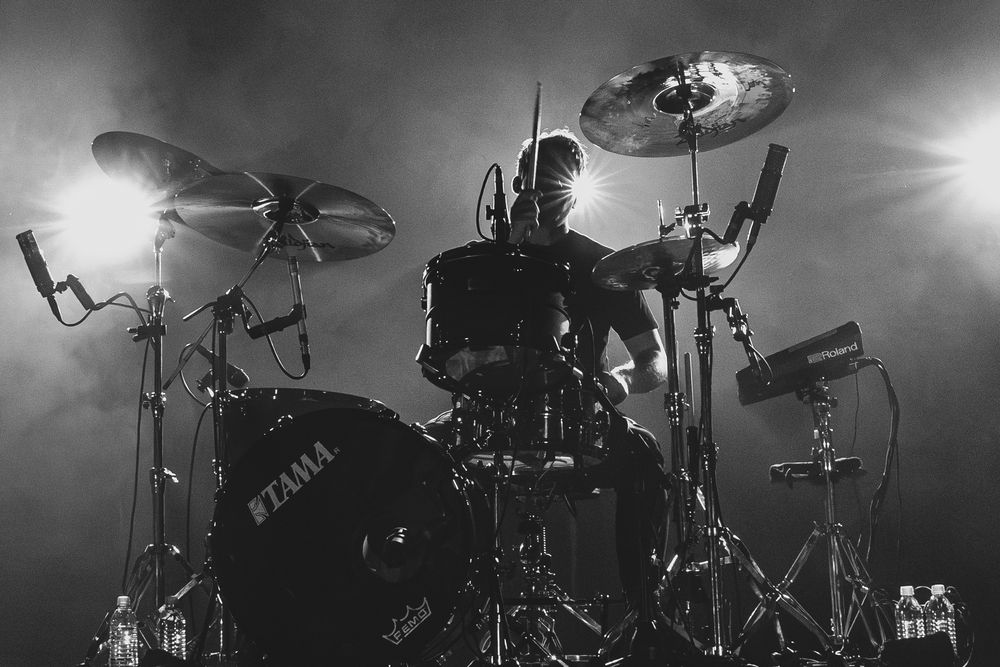Silhouetted drummer performing behind a Tama drum kit with cymbals and microphones under dramatic stage lighting in black and white.