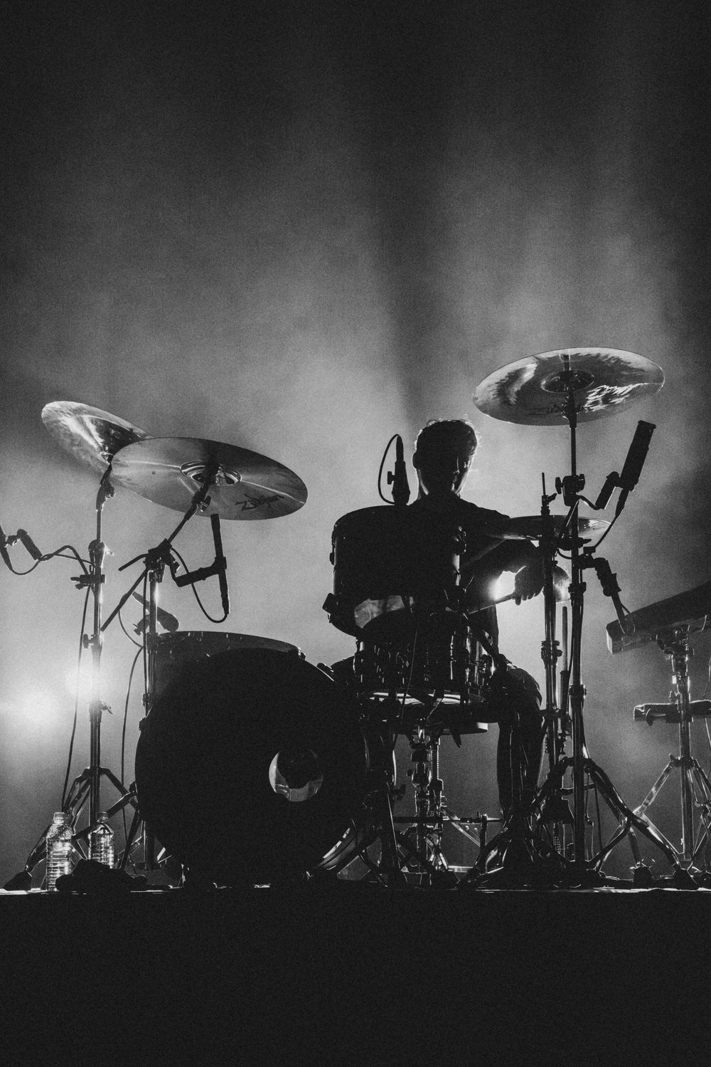 Silhouette of a drummer performing behind a drum kit with cymbals and microphones, backlit by dramatic stage lights in black and white.