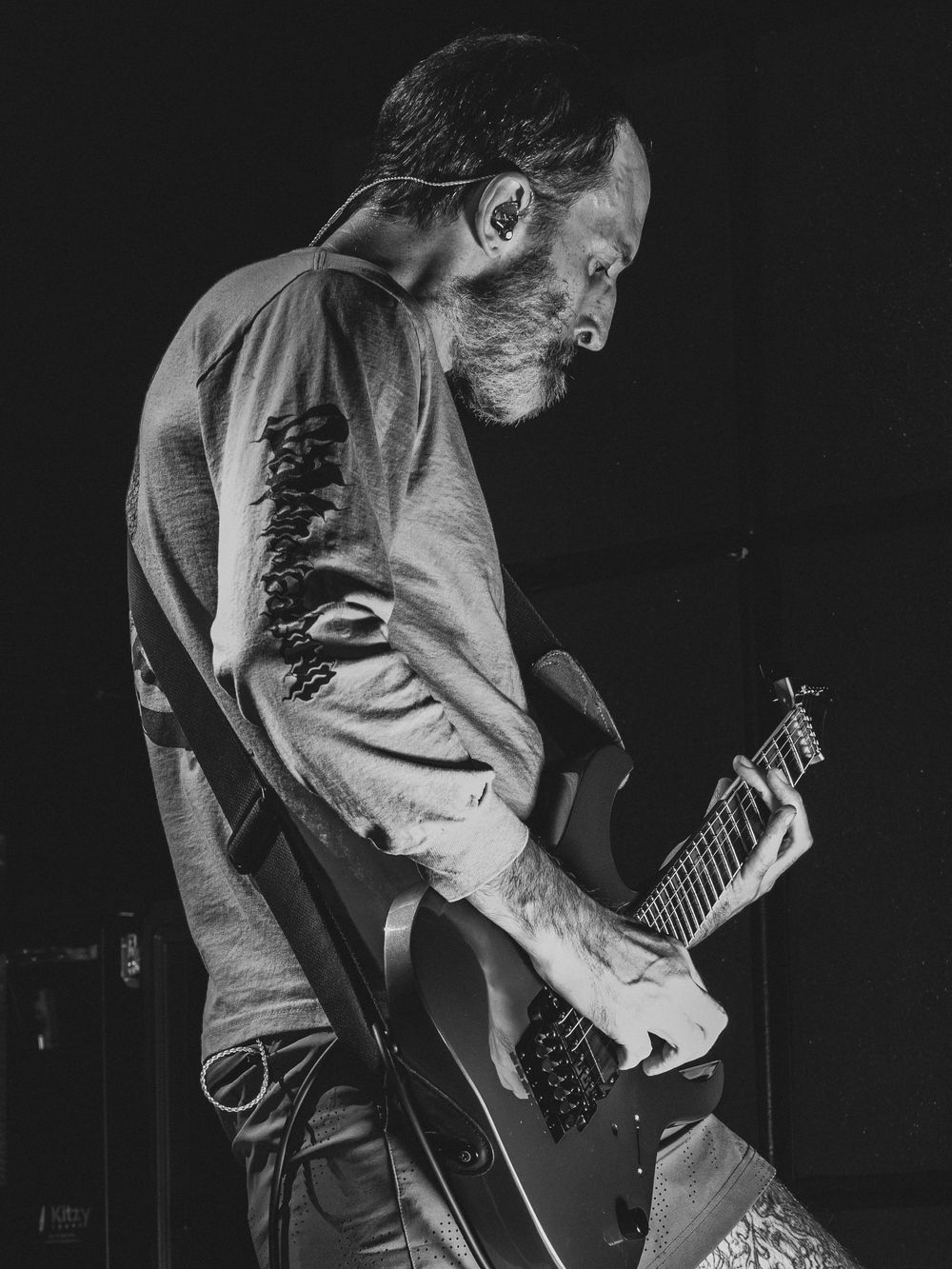 Black and white photo of a bearded guitarist in profile playing an electric guitar on a dark stage, wearing a graphic long-sleeve shirt.