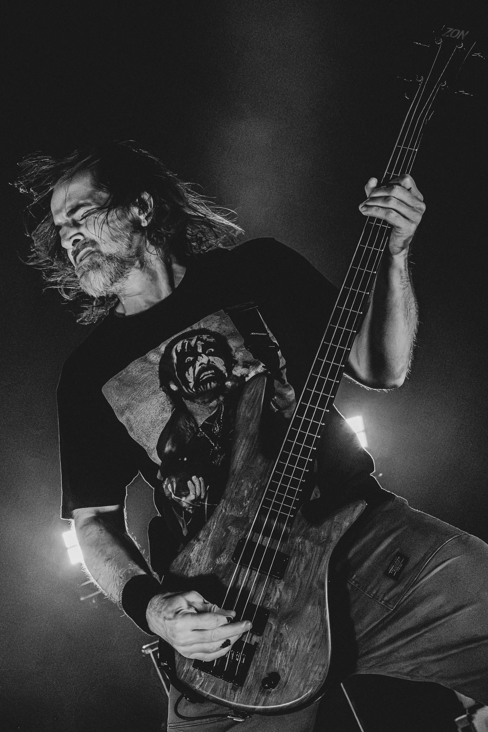 Black and white photo of a long-haired, bearded bassist playing an electric bass guitar on stage in a graphic tee under dramatic lighting.