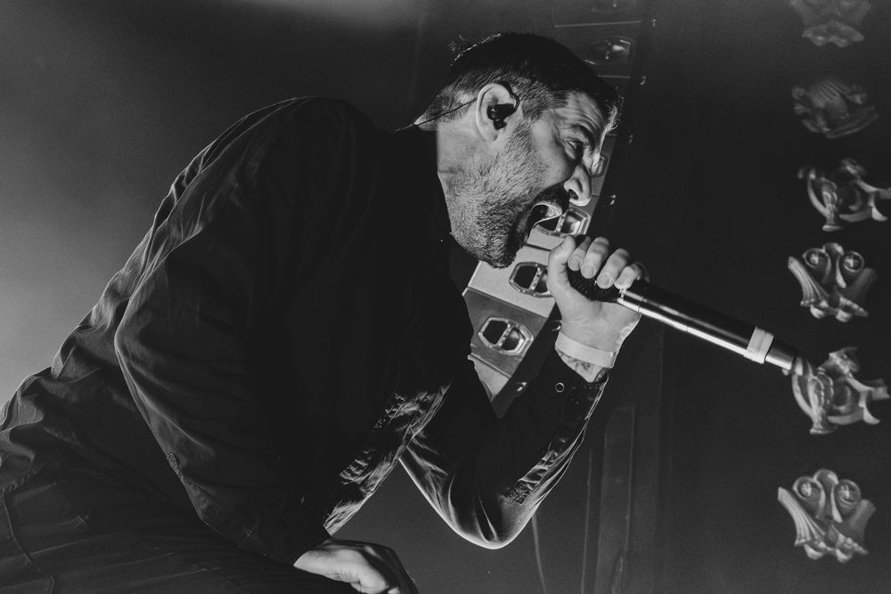 Black and white photo of a male vocalist singing intensely into a microphone on stage, with ornate decorative panels in the background.