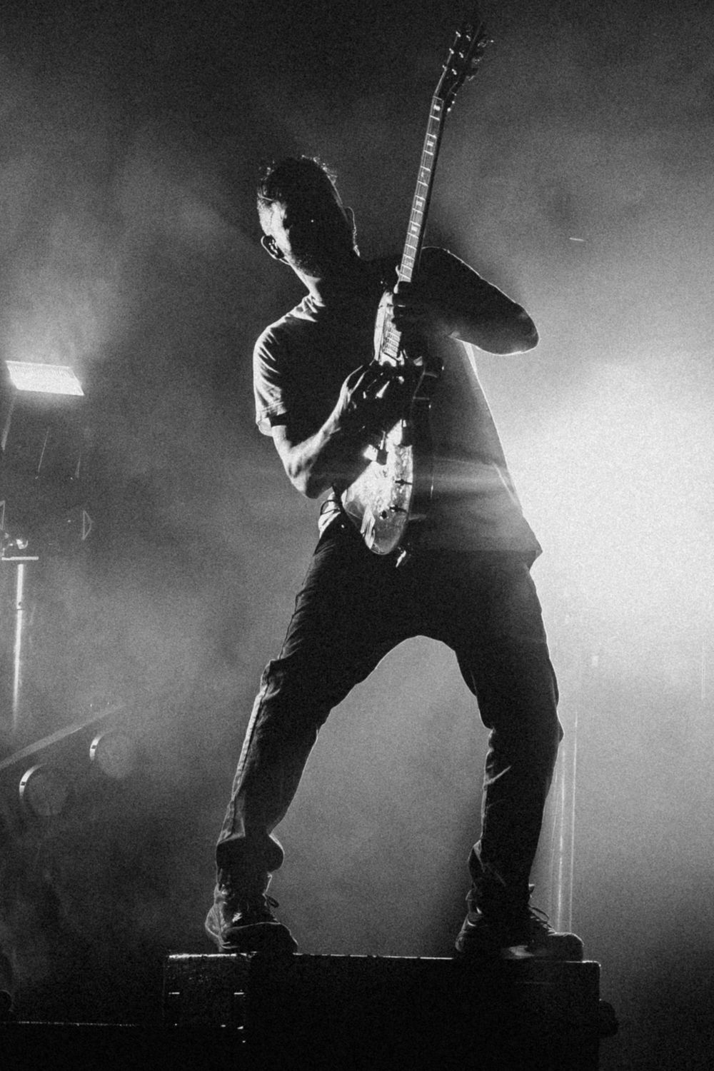 Black and white photo of a guitarist standing on a stage riser, playing an electric guitar amid hazy stage lights and smoke.