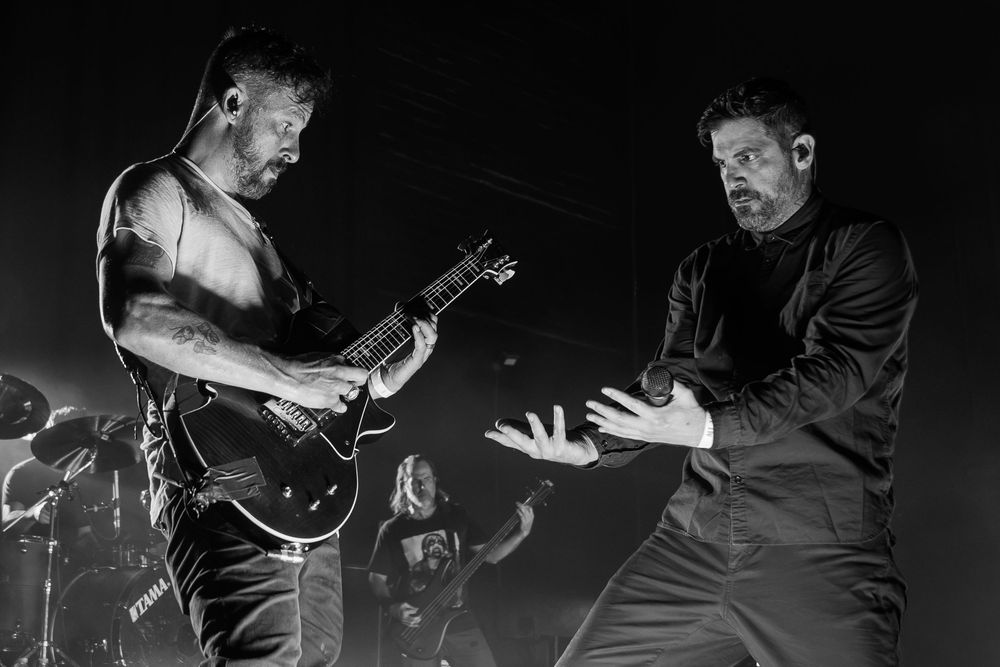 Black and white concert photo of a shirtless guitarist and a vocalist with outstretched hands performing on stage with a drummer behind.