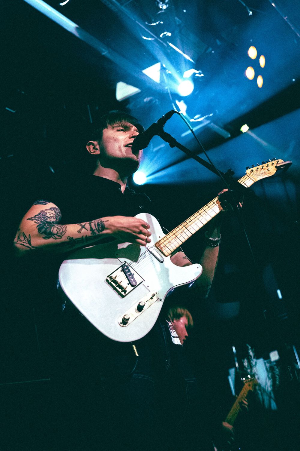 Tattooed guitarist sings into microphone while playing white Telecaster guitar under blue stage lights.