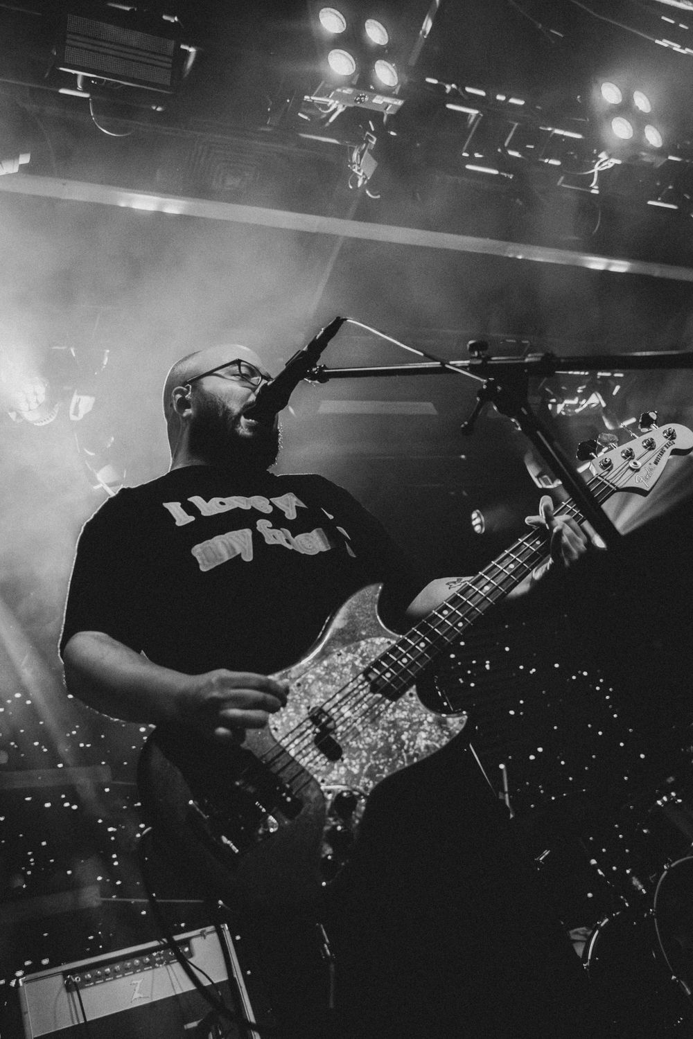 Bassist with glasses sings into microphone while playing bass guitar under smoky stage lights.