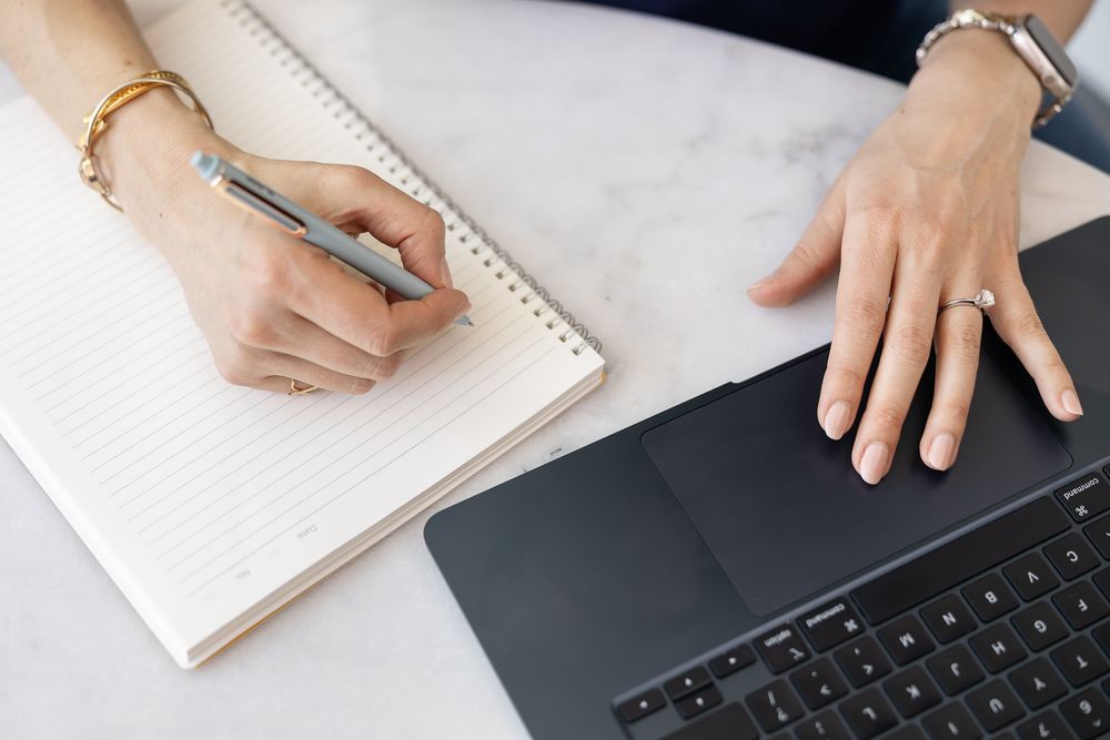 woman business owner sitting at desk taking notes in notebook with other hand on laptop computer for branded stock photo in Seattle, WA