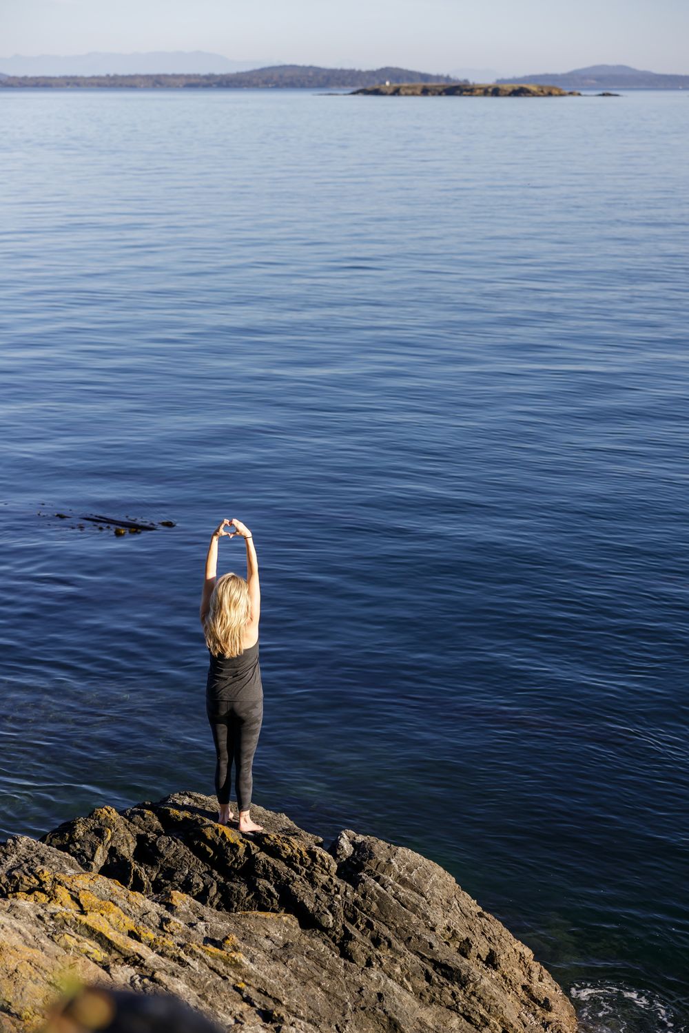 woman health coach standing on rocks by ocean with hands above head for lifestyle branded stock photo on Orcas Island, WA