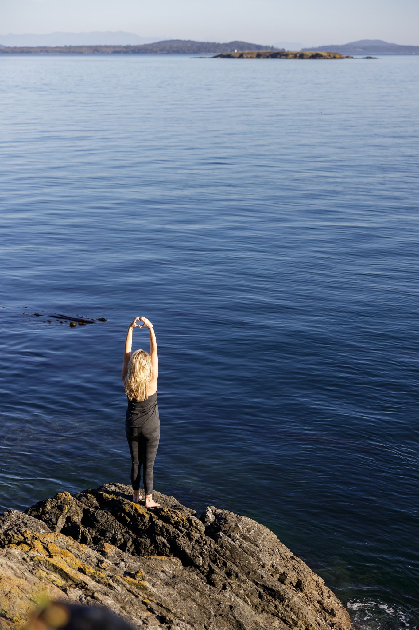 woman health coach standing on rocks by ocean with hands above head for lifestyle branded stock photo on Orcas Island, WA