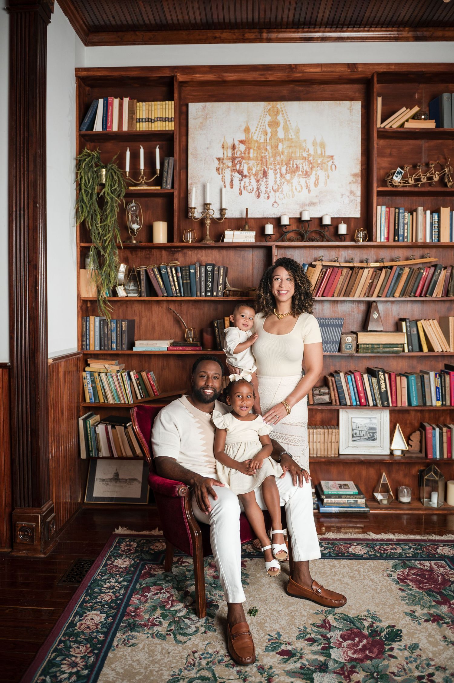 Lifestyle family portrait in cozy library room with wooden bookshelves and chandelier.