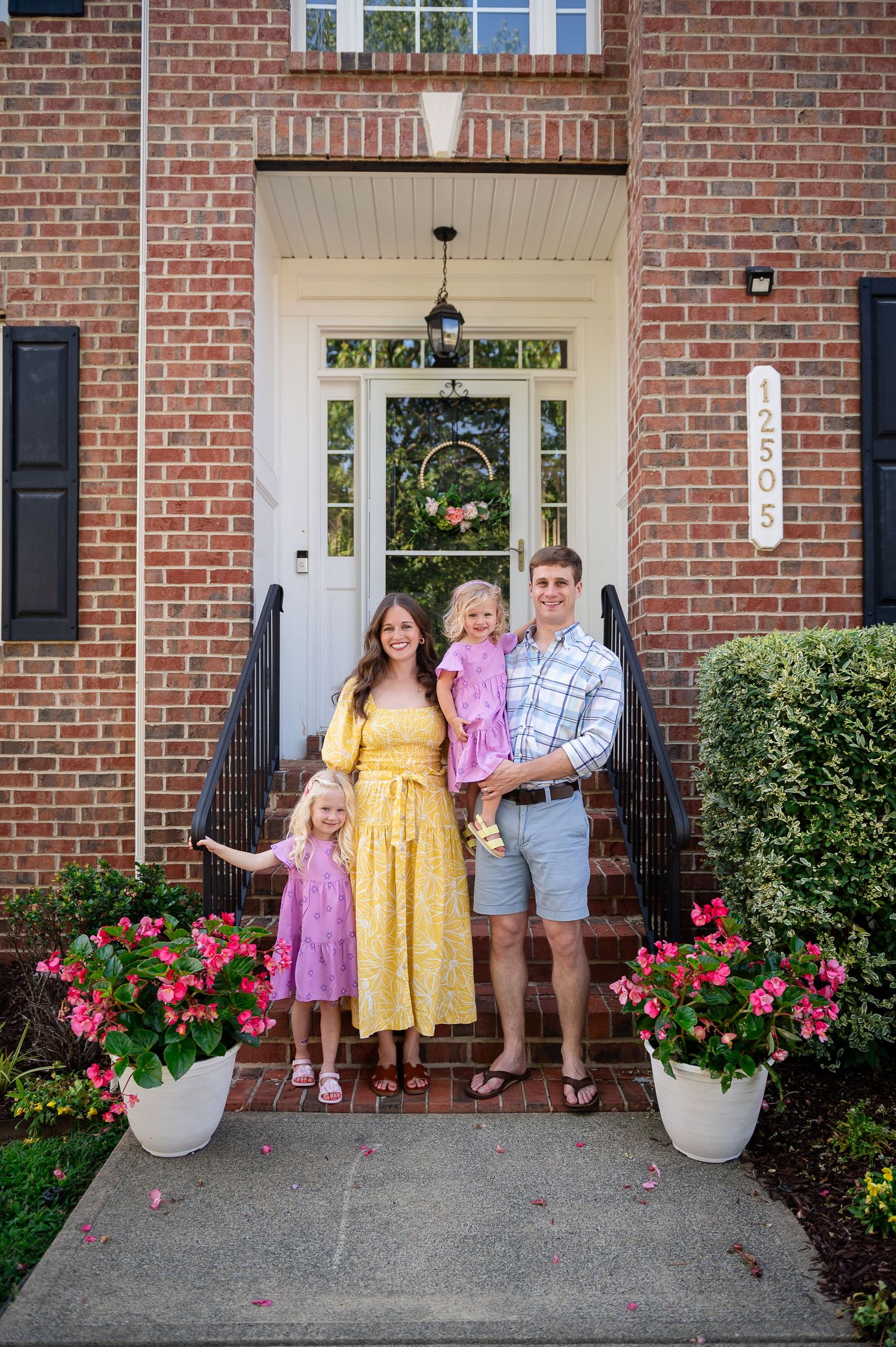 Family standing on brick house front porch with floral wreath and pink flowers.