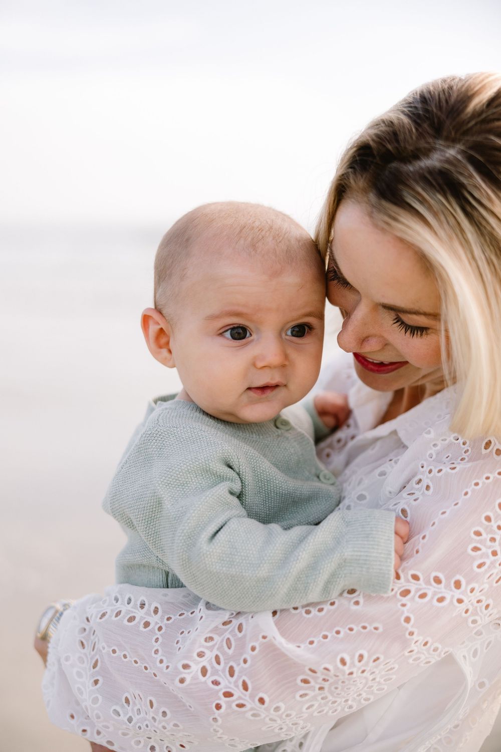 An intimate moment between parent and baby in white eyelet dress against light background.