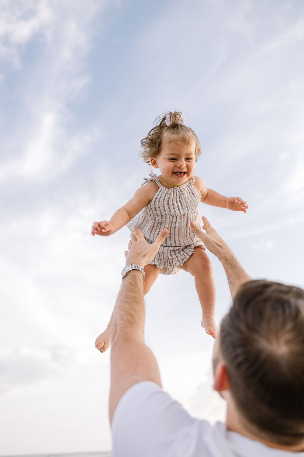 A baby in a knit romper is lifted joyfully into the air against a bright sky background.