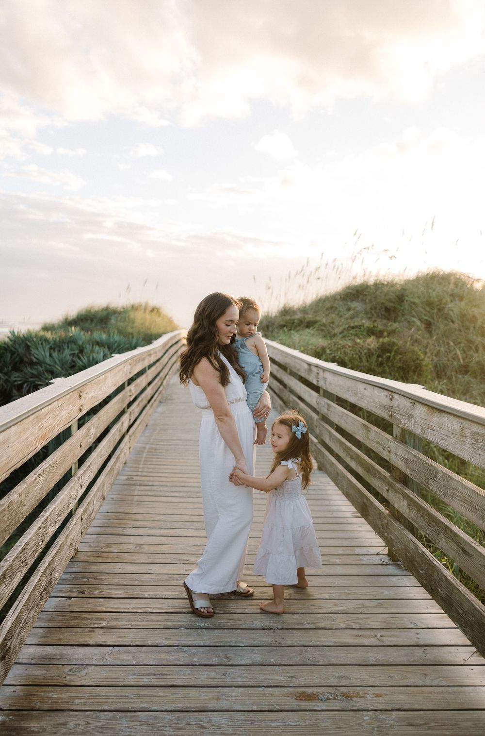 A parent and child in white dresses share tender moments while walking along a wooden beach boardwalk at sunset.
