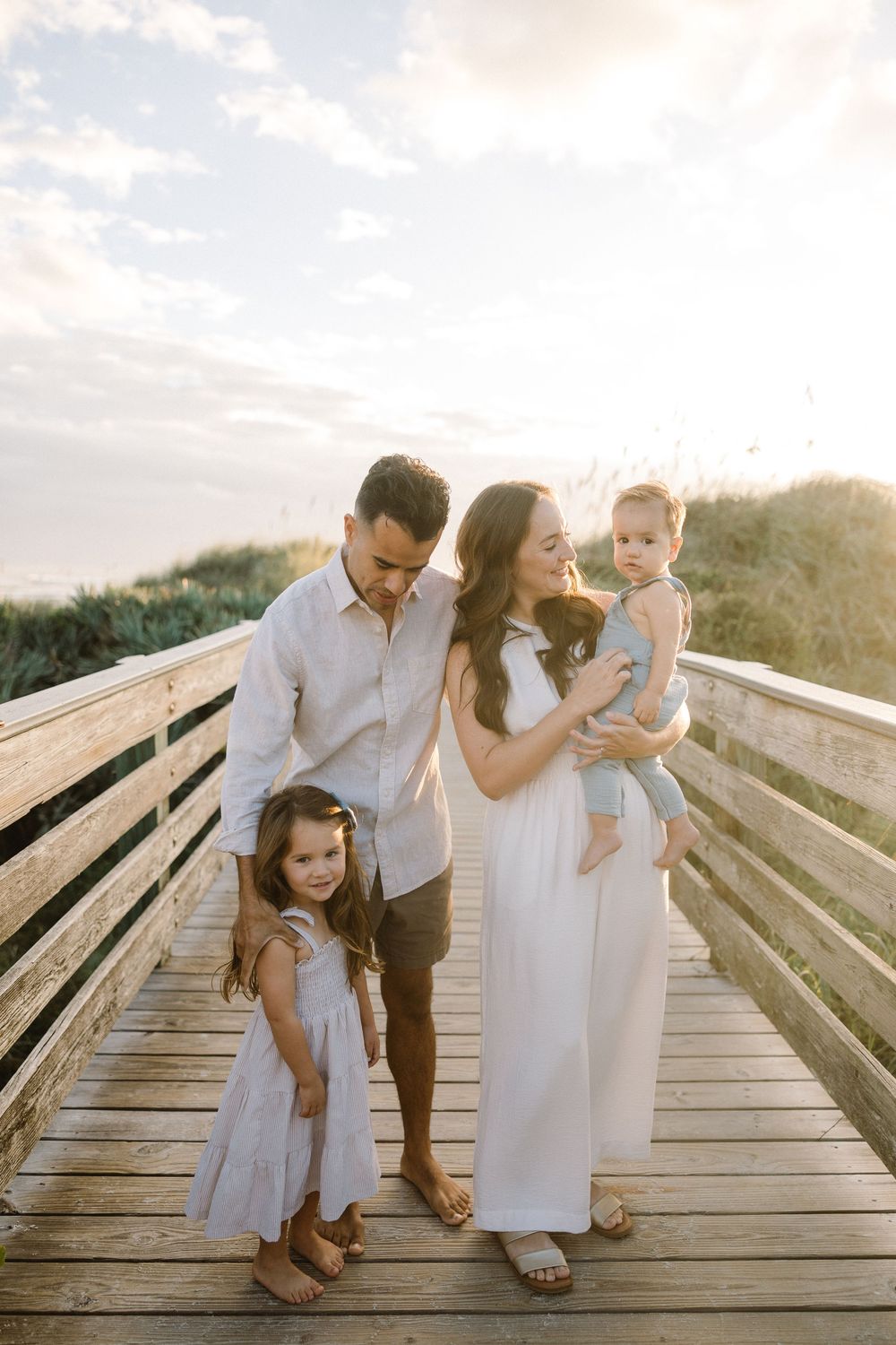 A family in white outfits shares tender moments together on a wooden boardwalk during sunset.