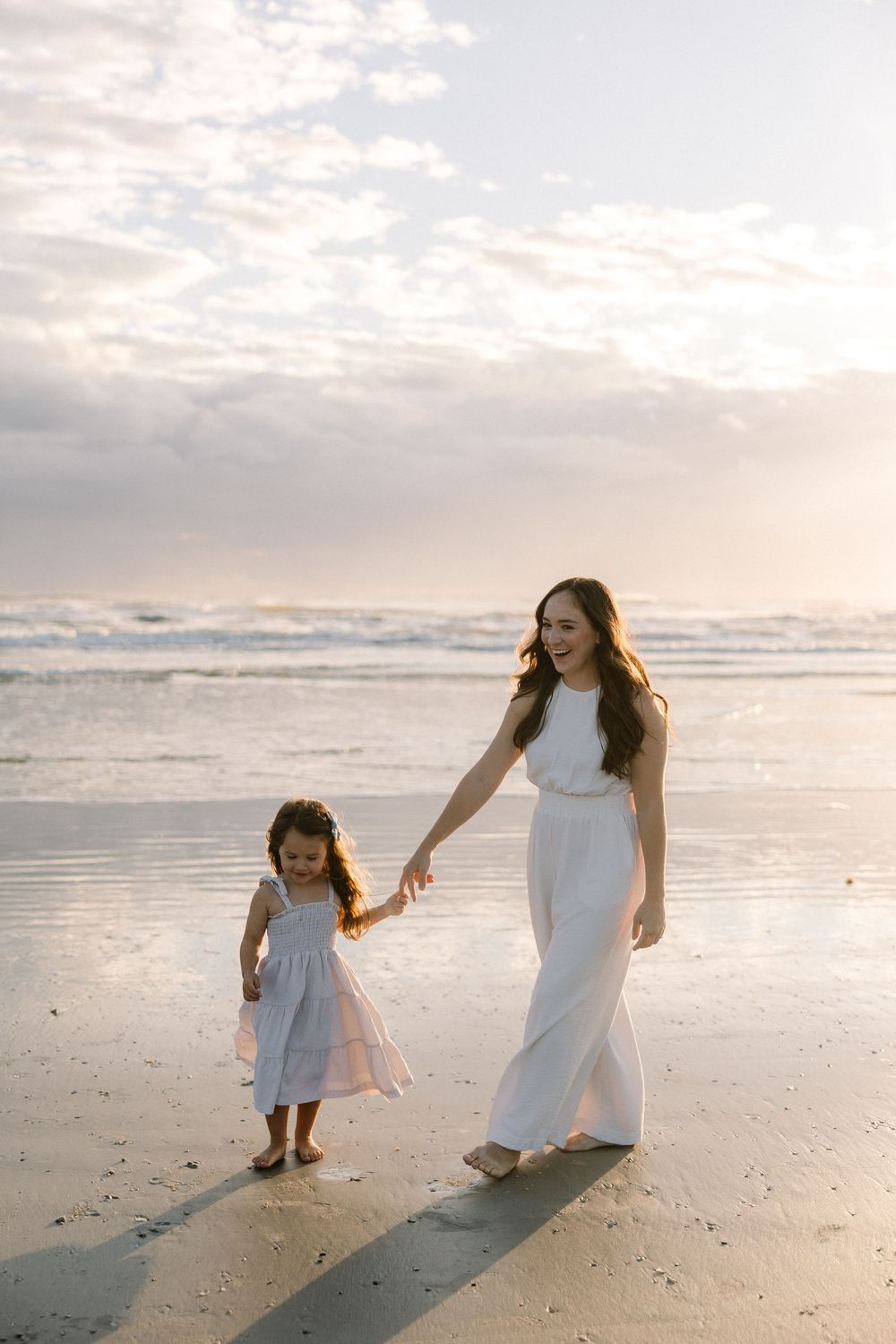 Mother and daughter in white dresses hold hands while walking along a peaceful beach at sunset.