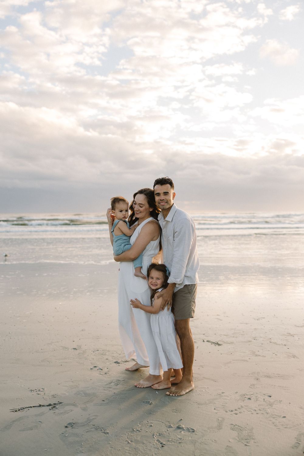 A family embraces together on a serene beach at sunset with waves and cloudy skies in the background.
