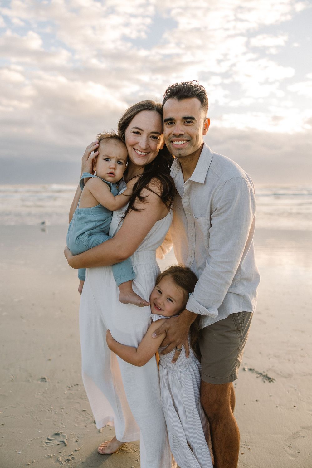 A family shares joyful moments together on a sandy beach at sunset wearing coordinated light blue and neutral outfits.