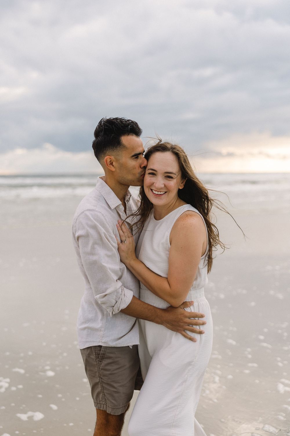 A couple shares joyful moments together on a beach during sunset with cloudy skies in the background.