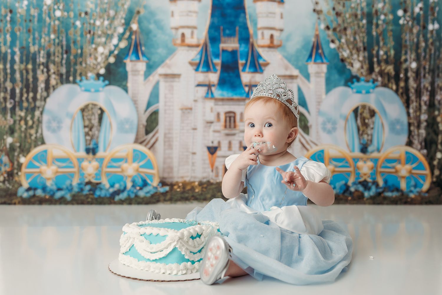 Baby in light blue dress sits on floor with birthday cake against winter wonderland backdrop with castle decorations.