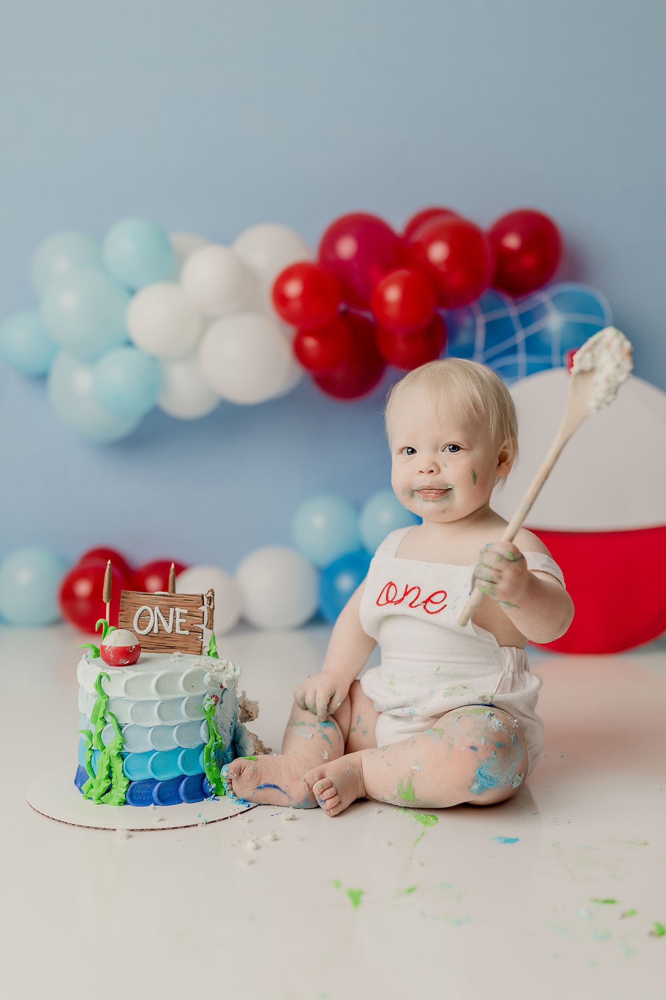 Colorful first birthday cake smash setup with red white and blue balloons and a decorated tier cake against blue backdrop.
