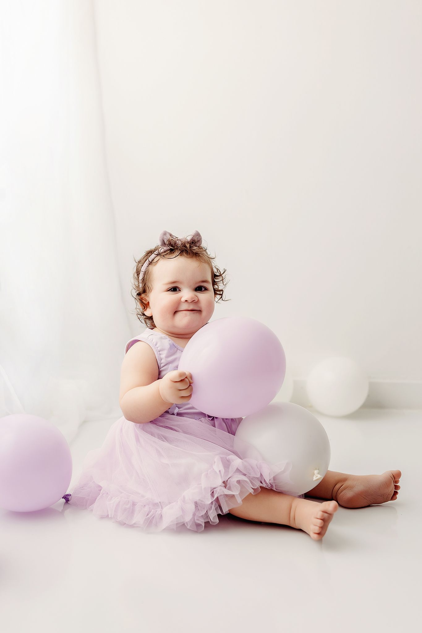 Baby wearing lavender tulle dress sits on white floor with pink and white balloons during portrait session.