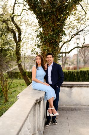 Engaged couple posing by stone balustrade with flowering ivy tree, woman in light blue dress and heels.
