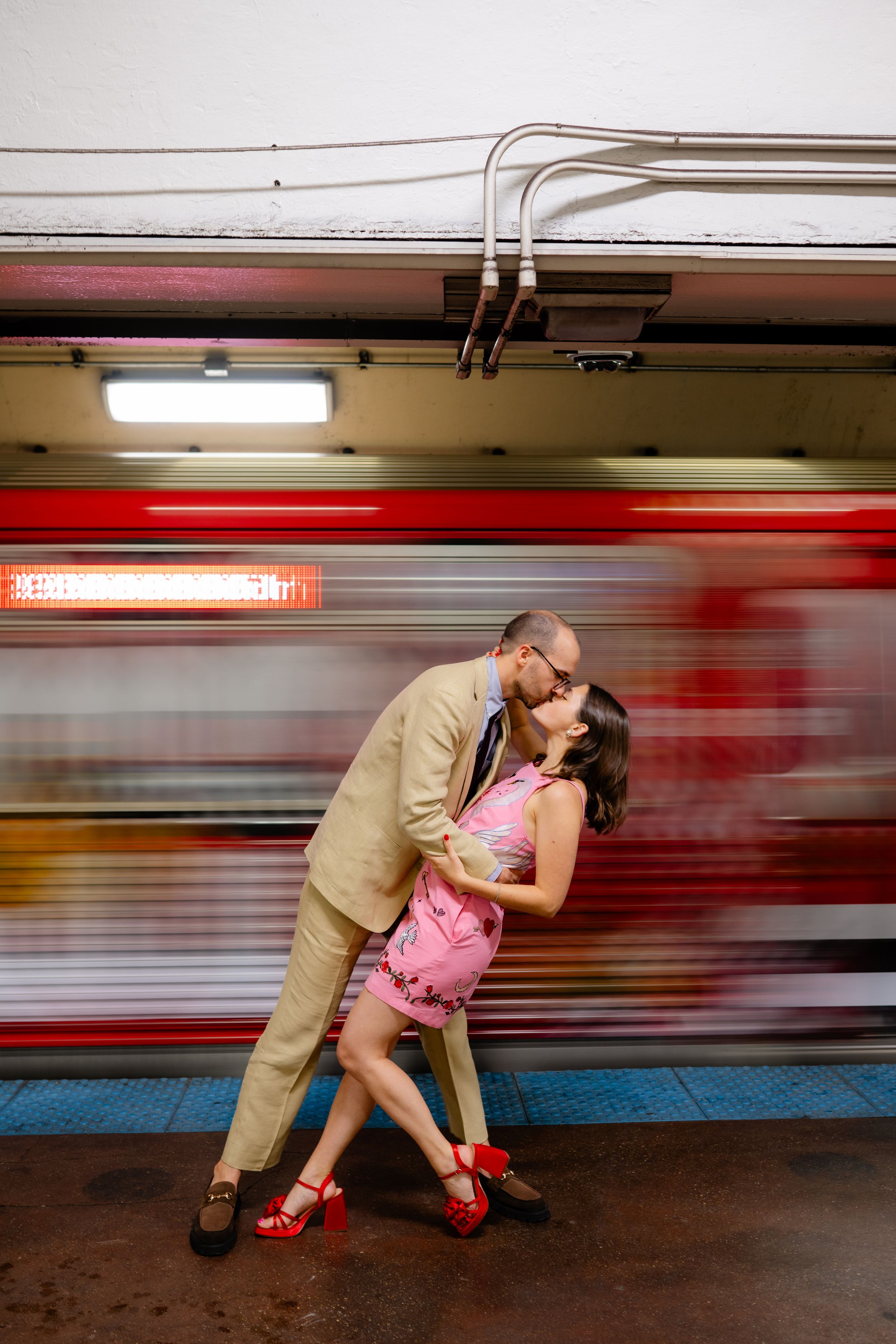 Couple sharing a romantic kiss on a subway platform as a red train blurs past behind them.