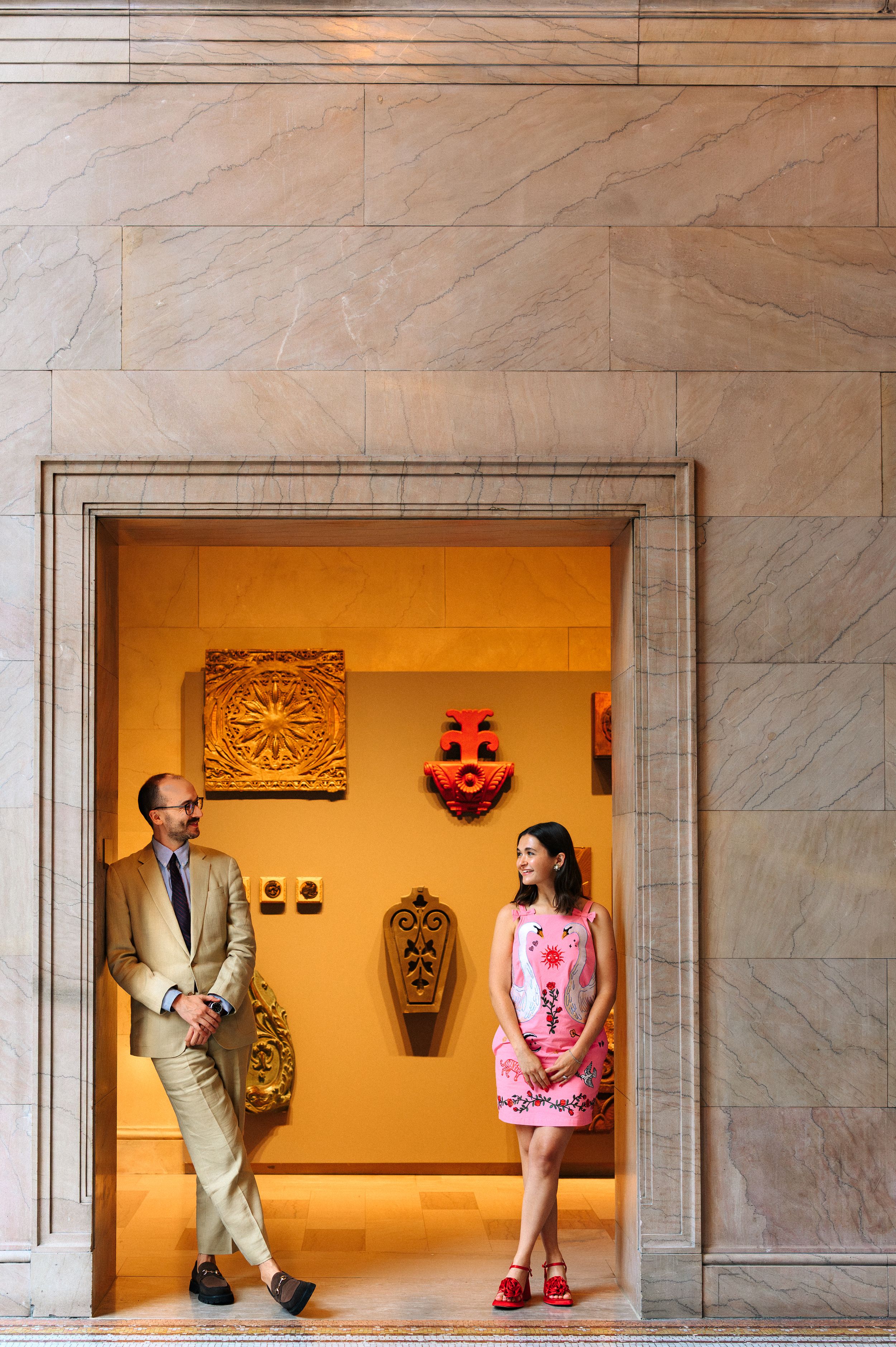 Man in tan suit and woman in pink dress stand in golden-walled museum alcove with decorative artifacts on display.