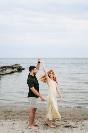 A couple dances together on a rocky beach shore with the ocean and cloudy sky in the background.