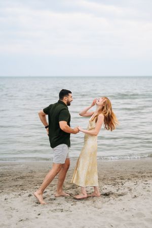 A couple dancing joyfully on a beach with the ocean in the background and cloudy sky above.