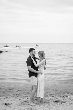 A romantic couple embraces on a beach at sunset with boats in the distance on the water.