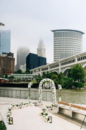 A wedding ceremony arch decorated with white flowers sits along the Cleveland waterfront with iconic city buildings in fog.