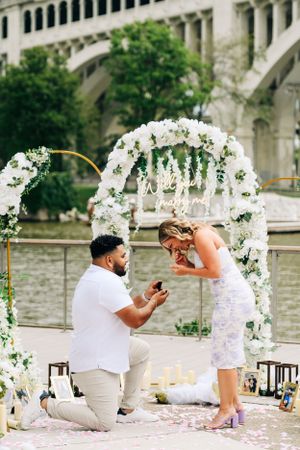 A romantic proposal scene under a floral white arch with petals scattered on the ground at an outdoor venue.