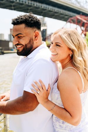 A happy couple embraces and laughs together by a waterfront while wearing white outfits on a sunny day.
