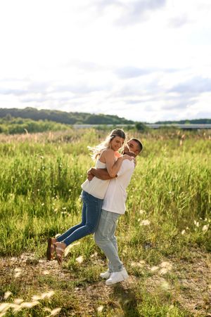 A joyful couple embraces and dances in a sunny green meadow, both wearing white tops and jeans.