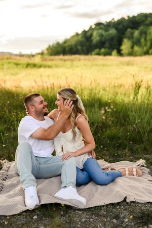Couple sitting on a blanket in a sunny field, smiling at each other in a tender romantic moment outdoors.