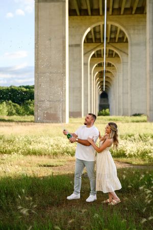 Couple celebrating under a bridge in a green field, man pouring champagne while woman in white dress smiles.