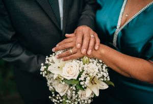 Hands with wedding rings rest on a delicate white rose and baby's breath bouquet against formal attire.